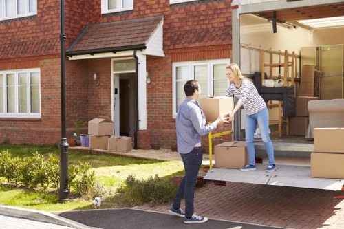 A scene outside a residential house in Catford shows a woman handing a cardboard box to a man standing on the driveway. The house has a brick exterior with white-framed windows and a small front garden with green grass and a lamp post. The garage door is open, revealing a cluttered interior with stacked cardboard boxes, a wooden shelving unit, and pieces of furniture including a sofa and a wardrobe, ready for moving. The man is dressed casually in a blue jacket and jeans, and the woman wears a striped top and light blue jeans. The scene captures the process of loading or unloading belongings during a house move, with the focus on collaboration and careful handling of household items. The overall environment appears bright with natural daylight, consistent with a typical residential street in London boroughs like Lewisham or Southwark near Catford. This image illustrates a professional house removal service, such as those offered by Catford Man with Van, facilitating safe and efficient relocations for local homeowners.