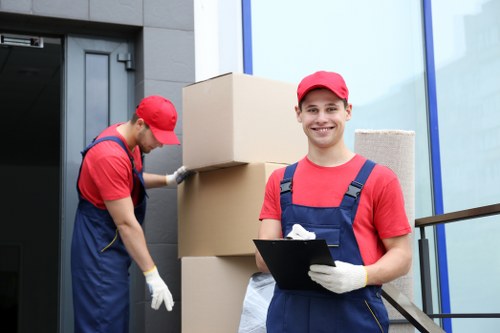 Two professional removals workers dressed in red caps, red shirts, and blue overalls are outside a modern building with large glass windows, preparing for a house move in Catford. The worker in the foreground is smiling at the camera, holding a clipboard and wearing gloves, while the other on the left is focused on adjusting or securing a stack of large cardboard moving boxes placed against the building's entrance. The boxes vary in size and are plain brown, with some partially opened or closed. The setting appears to be on a residential street, with the building's exterior made of dark grey brick or cladding. The environment is well-lit with daylight, and the overall scene depicts a professional house removal service in progress, illustrating typical activities involved in domestic relocations in the Catford area, London postcode SE6. This image emphasizes organized, careful packing and removal operations by Catford Man with Van, serving local household moving needs.