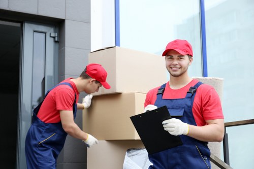 The image shows two male removal workers dressed in red caps and red shirts with navy blue overalls, engaged in a house moving task outside a modern residential building. The worker on the left is focused on adjusting or securing a large cardboard box, which is stacked on top of other boxes, all of which appear to contain household items. The worker on the right is smiling at the camera while holding a black clipboard, wearing white gloves, and standing near the bottom of the stacked boxes. The background features a contemporary building facade with glass windows and dark grey exterior walls, indicating a clean and professional environment typical of urban house removals in the Catford area, possibly within postcode SE6. The scene captures the process of packing or transporting household possessions, with emphasis on the equipment, such as the sturdy cardboard boxes, and the workers’ uniforms designed for a professional and organised removal service. The lighting suggests daytime, highlighting the organized and efficient approach typical of Catford Man with Van's house removal services, demonstrating careful handling of household belongings during a residential move in south-east London.