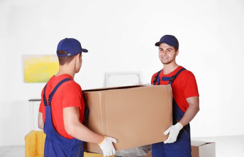Two removal workers wearing red T-shirts, blue overalls, and navy caps are lifting a medium-sized cardboard box together inside a bright, minimalistic room with white walls. The worker on the right is smiling and looking at the camera, wearing white gloves, while the other has his back turned. The room features a small yellow upholstered stool or piece of furniture in the background, and a white wall-mounted artwork with yellow and white hues. The scene suggests a professional house removal process, possibly in Catford, with a focus on careful handling of household items during a move. The lighting is natural and evenly distributed, highlighting the clean, uncluttered interior environment typical of a residential space where Catford Man with Van offers its removals services. Visible items include the cardboard box, the workers' uniforms, and part of the indoor setting, emphasizing the professional nature of the moving process.