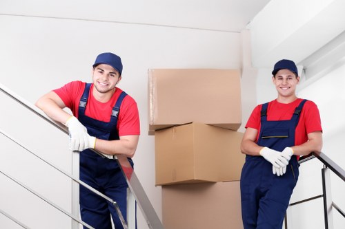 Two young male movers wearing red shirts, navy blue overalls, and dark caps are positioned on a staircase inside a residential property, preparing to load or unload cardboard boxes. The mover on the left is smiling and leaning on the metal handrail with his left arm, holding onto a metal pole, while the other stands on the right, also smiling, with his arms crossed in front of him. Behind them, there are two large, neatly stacked cardboard moving boxes, one slightly larger than the other, with the scene lit by natural indoor light. The environment appears to be a modern house with white walls and a staircase featuring a simple metal railing, suggesting a house or flat interior in a UK residential area. The movers are wearing protective gloves and casual work attire suitable for house removals, supporting the service offered by Catford Man with Van in the London area. The image conveys professionalism and readiness for a house move or relocation service within the local community, possibly near Catford in southeast London.