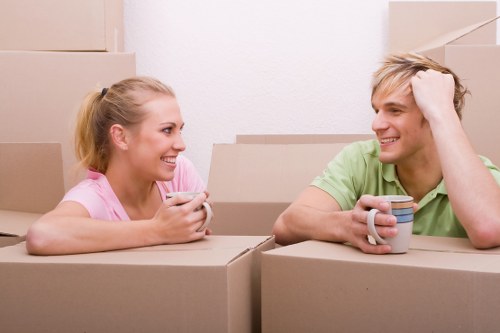 The image depicts a young couple, a woman with blonde hair tied back wearing a pink top and a man with blonde hair wearing a light green t-shirt, sitting inside a room filled with cardboard moving boxes. The woman is holding a smartphone and smiling, while the man is holding a striped ceramic mug and smiling back. The background shows a plain white wall and several stacked cardboard boxes, indicating they are preparing for a house move, possibly in the Catford area or nearby in South London. The scene is well-lit with natural or soft indoor lighting, emphasizing their happy expressions amid the clutter of packed household items, including cardboard boxes with visible textures of brown corrugated cardboard, hinting at an ongoing domestic relocation process undertaken by a professional removals service like Catford Man with Van. The setting suggests a residential space, potentially an interior hallway or living room, and the focus remains on the casual, positive interaction between the couple surrounded by their packed belongings in preparation for moving house.