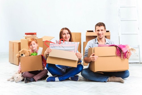 A family of three sitting on the floor of a bright, minimally decorated room with light grey walls and wooden flooring, surrounded by numerous cardboard boxes of varying sizes, some filled with household items and others sealed or partly open. The young girl on the left holds a small open box containing a plush teddy bear and a few toys, with her long hair and colorful clothing visible. In the center, a woman with long, brown hair is smiling and appears to be placing or taking an item from an open box, which contains wrapping paper or similar material, indicating packing activity. To her right, a man with short dark hair is holding a medium-sized cardboard box with a pink and purple scarf draped over its edge. Behind them are additional closed boxes stacked against the wall, suggesting an ongoing moving or packing process typical of a house removal in the Catford or nearby South London area, highlighting the families' preparations for relocation within the local postcode region.
