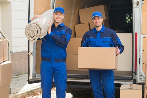 Two male movers dressed in blue work uniforms and caps are standing outside a residential property next to a parked removals van with its back doors open. One mover on the left is smiling and holding a rolled-up area rug with a textured, beige fabric and a spiral pattern, resting it on his shoulder. The other mover on the right is also smiling and holding a plain cardboard box at waist height. Behind them, the interior of the van is visible, filled with neatly stacked brown cardboard boxes of various sizes, some with visible flaps, and larger pieces of household furniture. The background shows part of a residential street with a house wall, a white garage door, and a small driveway. The scene depicts the process of house removals in Lewisham or the Catford area, with a focus on professional, careful handling of household belongings during a move. The lighting appears natural, suggesting a clear day, emphasizing the organized and prepared nature of the moving service provided by Catford Man with Van.