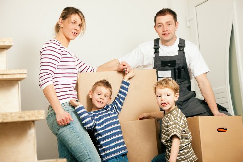 A family of four, including a woman with blonde hair wearing a red and white striped long-sleeve shirt and three young children, are seen in the interior of a residential home with light-colored walls and a staircase visible on the left. They are surrounded by opened cardboard boxes, some of which contain household items; the children are smiling and appear to be happy, with one boy wearing a blue and white striped shirt reaching into a box, another boy in a brown and beige striped shirt looking towards the camera, and a girl with long blonde hair partially visible behind a box. A male removal specialist from Catford Man with Van, dressed in a white shirt and black work overalls, stands behind the family, assisting with the moving process. The scene is well-lit, capturing the typical environment of a house during a residential move in the Catford area. The focus emphasizes the careful handling of household belongings during the house removal process.