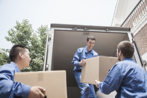 Three movers dressed in blue workwear unloading cardboard boxes from a removals van parked outside a residential property in Catford, London. The van's rear doors are open, revealing an interior space where two movers are actively carrying large, uniformly-sized cardboard boxes, while the third stands inside the van handing down a box to one of his colleagues. The boxes are sealed with packaging tape, and their surfaces are plain, indicating packed personal or household belongings. In the background, there are green trees and a residential building with a brick exterior and a balcony; the sky appears cloudy, casting diffused natural light on the scene. The environment suggests a professional house removals operation, with attention to careful handling of items typically associated with domestic moves, capturing the efficiency and organization of Catford Man with Van services in South London.