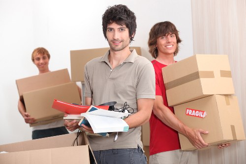 A group of three young men with light skin in a residential interior, likely a hallway or living room, preparing for a house move. The man in the foreground has dark, curly hair and is dressed in a light grey polo shirt; he is holding a stack of documents, a red clipboard, and a small pair of scissors, indicative of planning or organizational tasks. Behind him to the right, another man with curly brown hair, wearing a red shirt, is smiling while carrying two medium-sized cardboard boxes, one labeled 'FRAGILE' with clear brown packing tape. To the left, a third man with lighter hair, dressed casually in a white t-shirt, is holding a larger cardboard box. All are standing in front of a plain, light-colored wall, with some boxes placed on the floor, suggesting they are in the process of packing or preparing to move house. The scene emphasizes the practical aspects of residential removals with visible household items and packing materials typical of a house relocation in the Catford or greater South London area. Natural lighting illuminates the scene, creating a neutral and professional atmosphere consistent with house removal and relocation services.