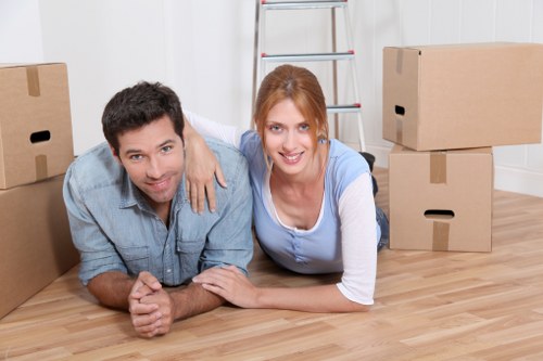 A couple of movers, a man and a woman, are lying on a light wooden floor inside a residential property, engaging in a move or packing activity. The man has dark hair and is wearing a light denim shirt, while the woman has red hair styled in a ponytail and is dressed in a blue and white top. They are smiling and looking directly at the camera, with the woman's arm draped over the man's shoulder. Surrounding them are several cardboard boxes of various sizes, some closed and others open, positioned in the foreground and background, indicating a packing or moving process. Behind them, there is a metallic step ladder leaning against a white paneled wall, and the room features a bright, clean atmosphere with natural lighting. The setting appears to be a typical interior of a house in the Catford area, consistent with house removals or moving service activities offered by Catford Man with Van, with the scene emphasizing the preparation phase of a home relocation within the local region of southeast London, specifically focusing on household items such as furniture and packing supplies.