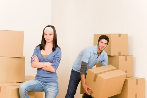 A man and a woman, both with light skin and brunette hair, are positioned in a room with plain white walls, surrounded by multiple cardboard boxes of various sizes, some taped shut with packing tape. The woman is sitting on a stack of boxes with her arms crossed, wearing a blue long-sleeved top and light blue jeans, with a neutral expression. The man is standing next to her, slightly bent forward, holding a large cardboard box and smiling, dressed in a light blue shirt layered over a darker long-sleeved shirt and dark trousers. The boxes are arranged in the foreground and background, suggesting a moving or packing process, consistent with house removals services. The lighting appears bright and even, typical of indoor residential settings, subtly indicating a home environment such as a living room or hallway in Catford or nearby London areas. The scene reflects the process of packing and preparing household items for relocation, relevant to Catford Man with Van's domestic removals services across the SE6 postcode area.