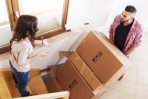 A man with a neatly trimmed beard and short dark hair, wearing a red plaid shirt over a black T-shirt, is seen carrying two large cardboard boxes, one stacked on top of the other, at the base of a staircase inside a residential property in Catford. The boxes display standard handling symbols and are made of brown corrugated cardboard with visible tape sealed along the edges. A woman with long, wavy brown hair, dressed in a beige top and blue jeans, stands on the staircase, gesturing towards the boxes, possibly giving instructions or discussing the move. The interior setting features a cream-colored wall and a large window with a wooden frame, letting in natural light, adjacent to a white cupboard or cabinet. The staircase is wooden with a light finish, and the floor nearby is tiled in a neutral tone, typical of a suburban house in SE6 postcode area. The scene depicts a typical house removal process, with focused activity around loading household belongings, consistent with the services offered by Catford Man with Van.