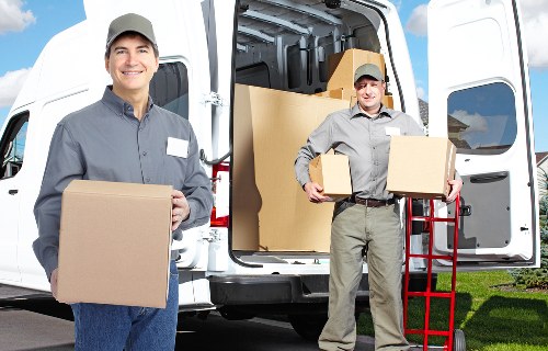 Two professional movers from Catford Man with Van are loading cardboard boxes into the rear of a white removals van parked in a residential area. The first mover, positioned on the left, is smiling and holding a medium-sized cardboard box, dressed in a grey uniform with a cap. The second mover, on the right, is carrying two boxes, one in each hand, and is also dressed in a grey uniform with a cap. The van's rear doors are open, revealing additional boxes stacked inside, and a red hand trolley is leaning against the side of the vehicle. The scene is set outdoors with green grass and a clear sky, indicating daytime. The focus is on the careful handling of household items such as cardboard boxes, which are used for moving personal belongings during house removals in the Catford area. The image conveys a professional and organized approach to residential moving services, relevant to families and individuals planning to relocate within or near London postcode areas like SE6 or SE4.