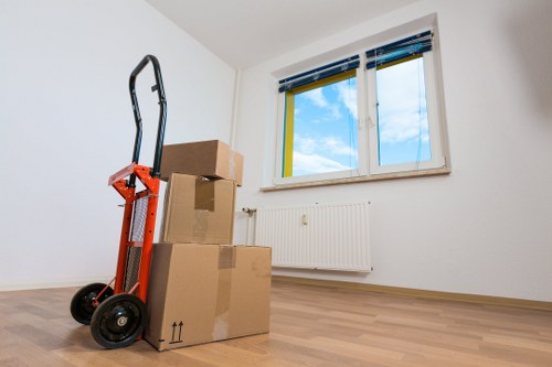 The image depicts a minimally furnished interior room, likely part of a house or flat in Catford, with light wooden flooring and plain white walls. In the foreground, a red hand truck with black wheels is leaning against a stack of three cardboard boxes in various sizes, some of which are sealed with packing tape. The boxes appear sturdy and are suitable for moving fragile or personal items. Behind the boxes, a white radiator is mounted beneath a window with a white frame, yellow and blue blinds, and a view of a partly cloudy sky outside. The room is well-lit by natural daylight coming through the window, creating a bright and neutral environment. The absence of furniture or clutter suggests that the space may be in the process of being prepared for a move or renovation. The scene reflects typical elements involved in residential removals, with attention to safe packing and stacking. The presence of the boxes and hand truck subtly highlights professional house removals and moving services, associated with companies like Catford Man with Van, serving local areas around postcode SE6 in South East London.