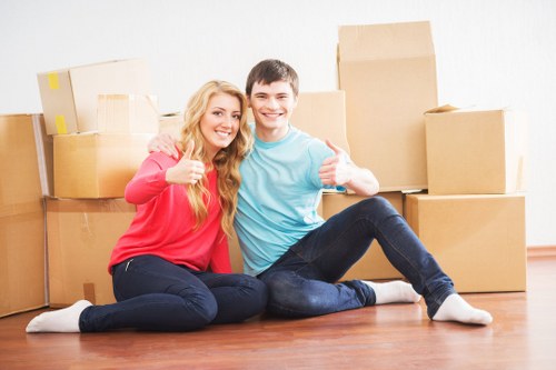 A young man and woman sit on a wooden floor in a residential interior, surrounded by several cardboard moving boxes of various sizes, some sealed with packing tape. The woman, wearing a red long-sleeved top and dark jeans, has long blonde hair and is giving a thumbs-up while smiling at the camera. The young man, dressed in a light blue T-shirt and dark jeans, has short dark hair and is also smiling with his arm around the woman’s shoulders, giving a thumbs-up. The background features a plain white wall, and natural light illuminates the scene, creating a bright and positive atmosphere indicative of a household relocation in the Catford or South East London area, consistent with house removals services. The scene emphasizes the process of packing and moving furniture and personal belongings, highlighting typical household items ready for transportation during a house move.