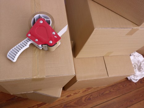 The image shows a close-up view of a cardboard box on a wooden floor, with a red and white handheld tape dispenser resting on top of it. The tape dispenser’s handle is white with a textured grip, and the red plastic housing covers the tape roll spindle. Several other cardboard boxes of varying sizes surround the main box, some stacked, indicating packing or moving activity. The scene appears to be inside a residential space, possibly a hallway or room set up for house removals, with the focus on packing materials used by Catford Man with Van specializing in local removals within the Lewisham area near Catford. The lighting is natural and evenly distributed, highlighting the textures of the cardboard, the wooden floor, and the tape dispenser, emphasizing a professional, organized environment related to house moving services.