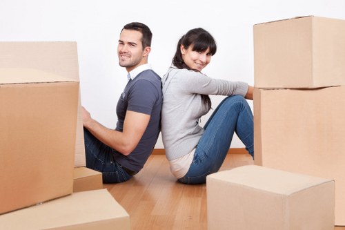 A young man and woman are seated on a light wooden floor inside a residential property, surrounded by several large cardboard moving boxes. The man, on the left, has dark hair, a short beard, and is dressed in a dark blue t-shirt and jeans, smiling gently towards the camera. The woman, on the right, has dark hair tied in a ponytail and is wearing a light grey long-sleeved top with blue jeans, sitting with her back against some boxes and smiling at the camera. The boxes vary in size, some stacked and others placed individually around them, with the setting appearing to be a bright, tidy room with white walls. The scene reflects a house moving or packing process, with the focus on the casual and organized atmosphere of a family or individuals preparing for a relocation. The company name, Catford Man with Van, is associated with local removals services in the London area, subtly suggested by the home environment and context.