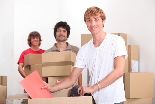 Three individuals with light skin and casual clothing are in a well-lit room filled with cardboard moving boxes, indicating a house removal or relocation scene in Catford. The young man in the foreground, wearing a white t-shirt, is smiling and holding a large pink envelope or document in his right hand, while resting his left arm on a partially opened cardboard box. Behind him, two other individuals, a woman in a red shirt and a man in a beige shirt, are also smiling and each holding or standing near stacked cardboard boxes of various sizes, some with labels attached. The background features a plain white wall, suggesting an indoor residential setting typical of a house or flat in the Catford area. The scene captures the active process of packing or unpacking, with the focus on household items such as boxes and personal belongings, highlighting the professional house removal services provided by Catford Man with Van.
