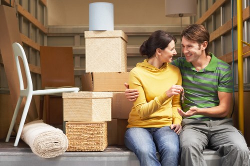 A young couple with light skin sitting inside a partially visible storage unit or moving truck, surrounded by packed household items including stacked cardboard boxes, a wicker basket, a rolled-up beige carpet, and a white plastic chair. The woman, wearing a yellow top and blue jeans, is smiling and embracing the man, who is dressed in a green and gray striped polo shirt and gray trousers. The background shows wooden shelves and a neutral wall, indicating a typical moving or storage environment in or around a residential property in the Catford area, related to house removals services by Catford Man with Van. The scene is well-lit with natural or artificial light, capturing an atmosphere of packing or preparing for a move.