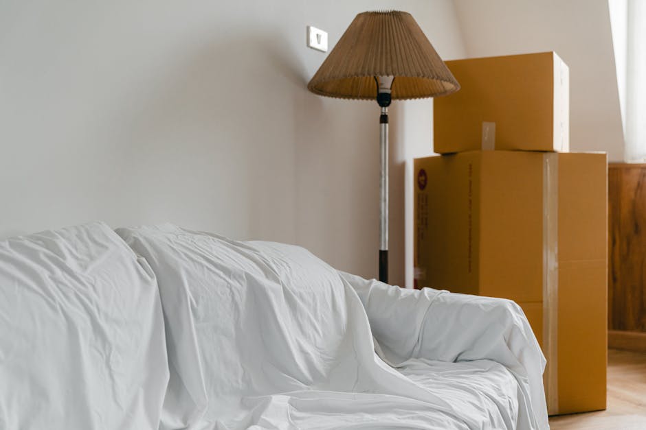 Inside a room during a home relocation process, a white sofa covered with a protective sheet is positioned in front of a plain light-colored wall, with a tall standing floor lamp featuring a brown ple